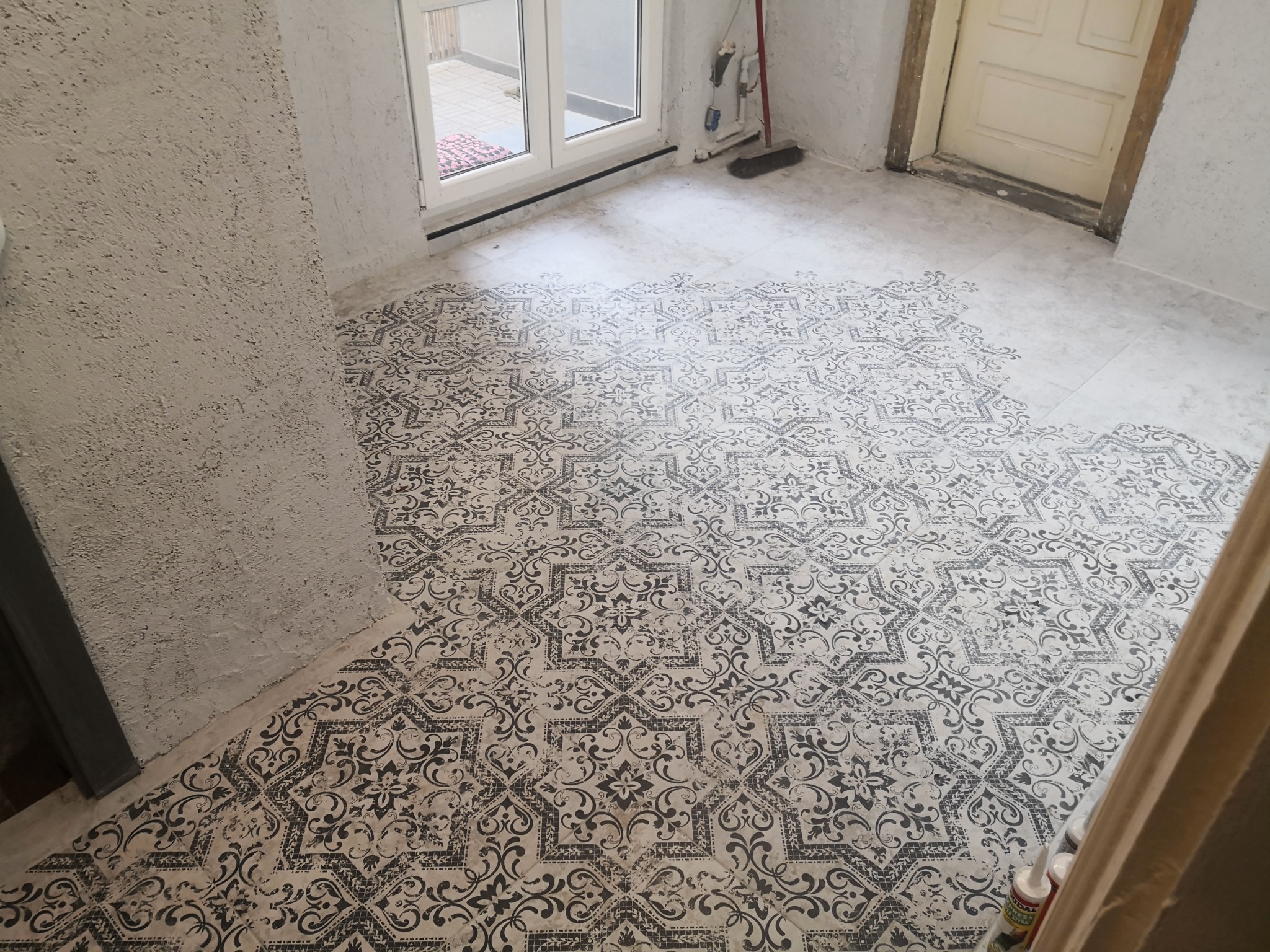 Fragment podłogi z ułożonymi wzorzystymi płytkami w odcieniach szarości i bieli, widoczne niedociągnięcia w wykończeniu przy ścianach i drzwiach, resztki zaprawy i narzędzia w tle.