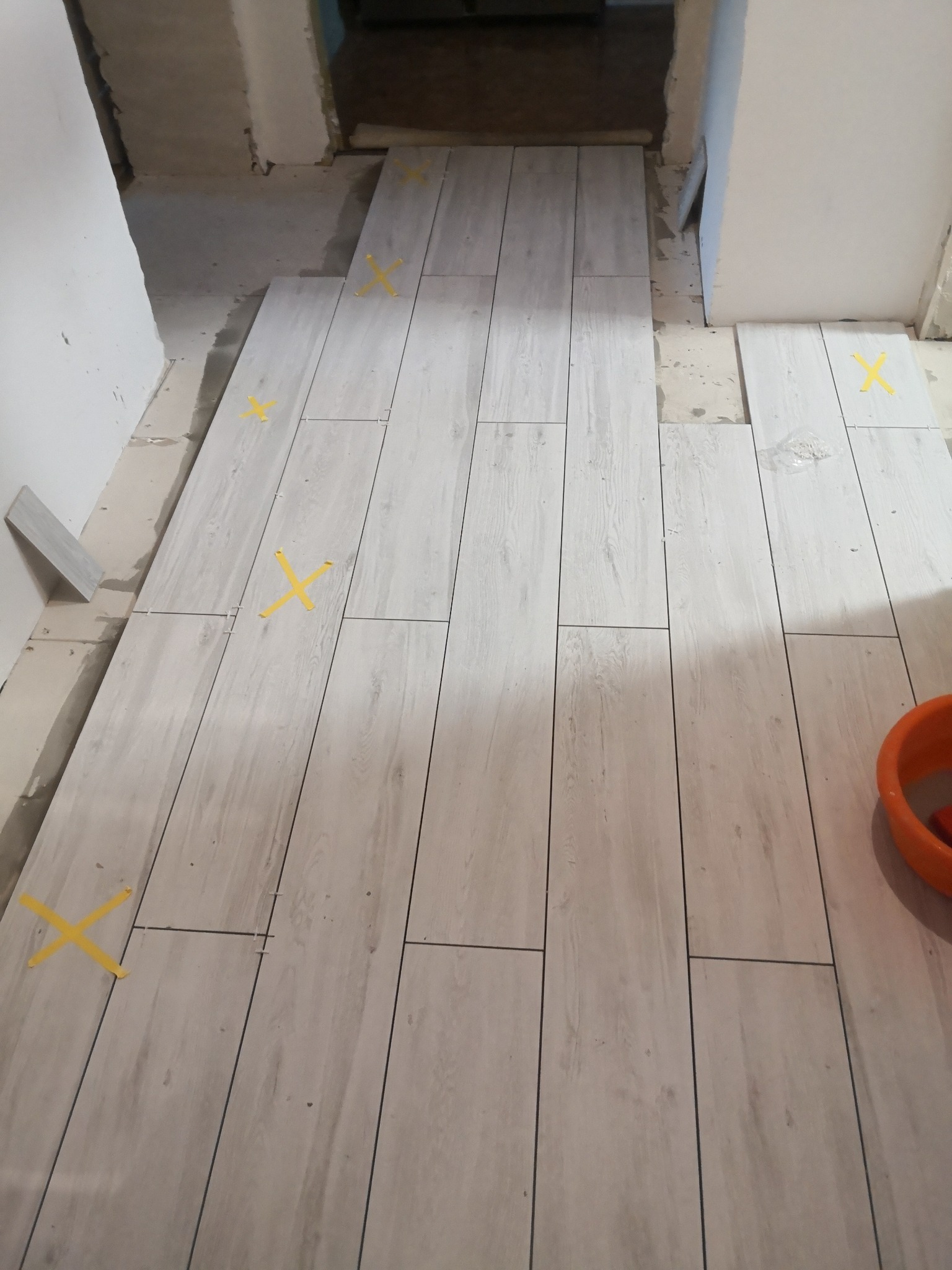 Układanie jasnych, podłużnych płytek imitujących drewno na podłodze w korytarzu, z żółtymi krzyżykami dystansowymi i widocznym pomarańczowym wiadrem w rogu.