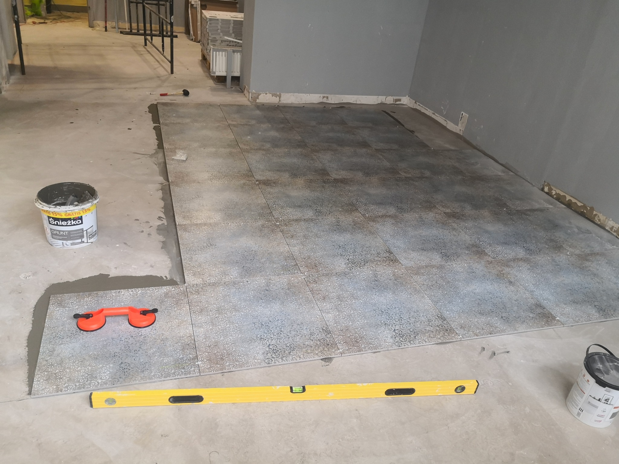 Układanie płytek podłogowych z wzorem, widoczna poziomica, przyssawka do płytek oraz wiadro z gruntem Śnieżka.