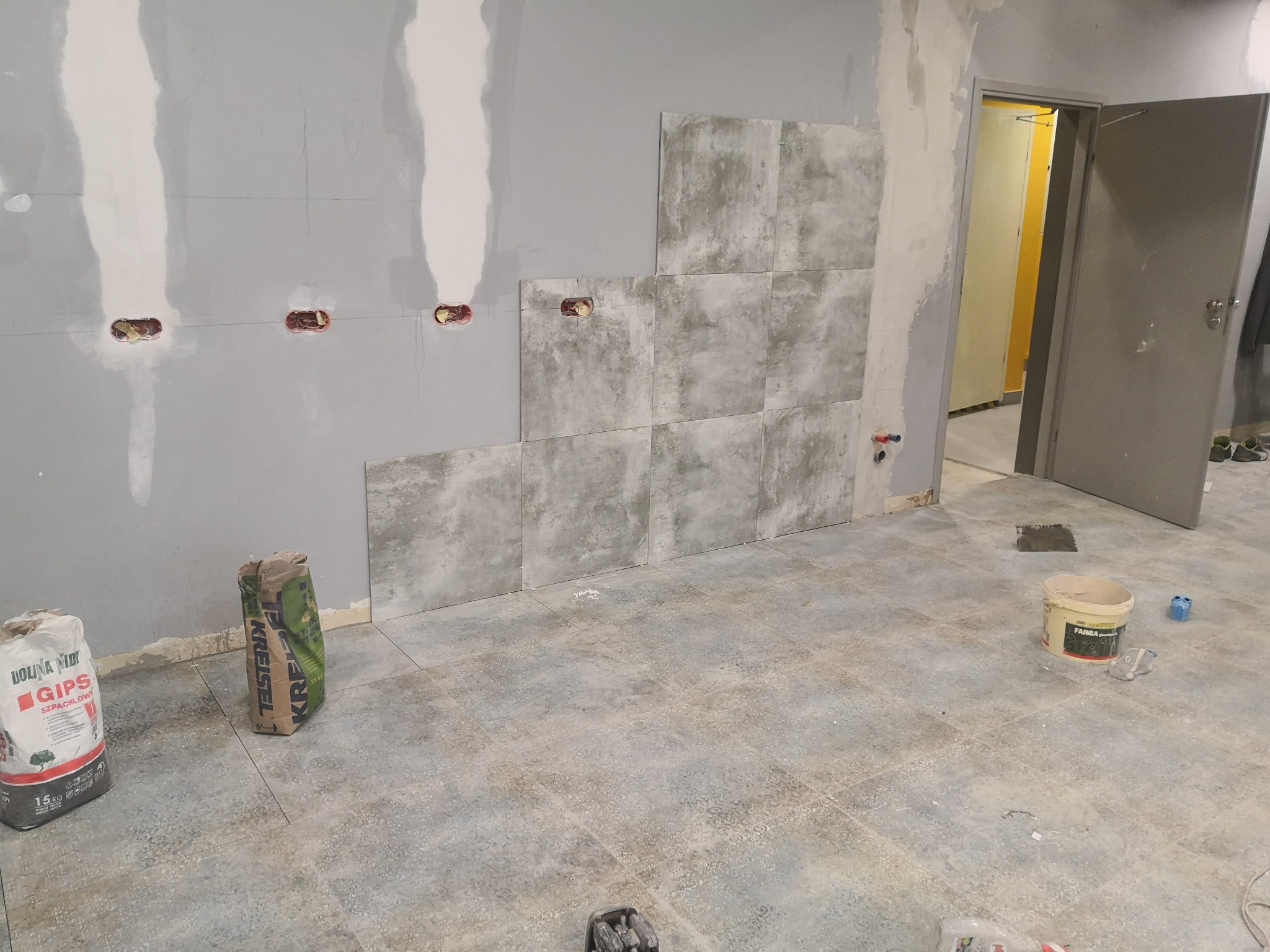Układanie szarych płytek imitujących kamień na ścianie z gładzią gipsową, widoczne puszki elektryczne, worki gipsu szpachlowego Dolina Nidy i kleju KREISEL, oraz wiadro po farbie Fabra