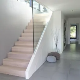 Jasne, drewniane schody z przezroczystą, szklaną balustradą w nowoczesnym wnętrzu z szarymi płytkami podłogowymi. W tle widoczny korytarz i dekoracyjne pufy. Minimalistyczny design.