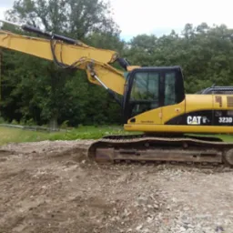 Żółta koparka gąsienicowa CAT 323D LVA na nierównym terenie z ziemią i kamieniami, na tle zieleni drzew.