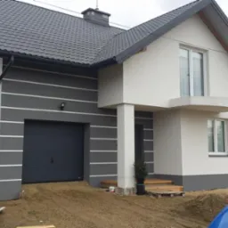 Nowoczesny dom jednorodzinny po renowacji elewacji, z szarą fasadą i garażem, białe elementy dekoracyjne, ciemna dachówka. Widok z zewnątrz.