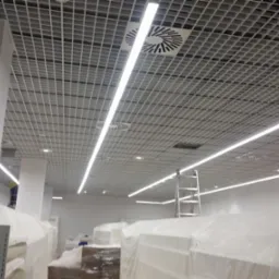 Sufit rastrowy z zamontowanym oświetleniem liniowym LED i kratkami wentylacyjnymi w nowym obiekcie, z widoczną drabiną i zabezpieczonymi folią elementami wyposażenia.