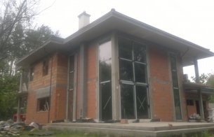 Niedokończony budynek z widocznymi przewodami elektrycznymi wystającymi ze ścian, przygotowanymi do podłączenia instalacji, z oknami zabezpieczonymi taśmą, w tle drzewa.