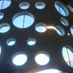 Ściana z licznymi okrągłymi otworami, przez które widać niebieskie niebo i fragment konstrukcji budynku, z widocznym żurawiem budowlanym w tle.