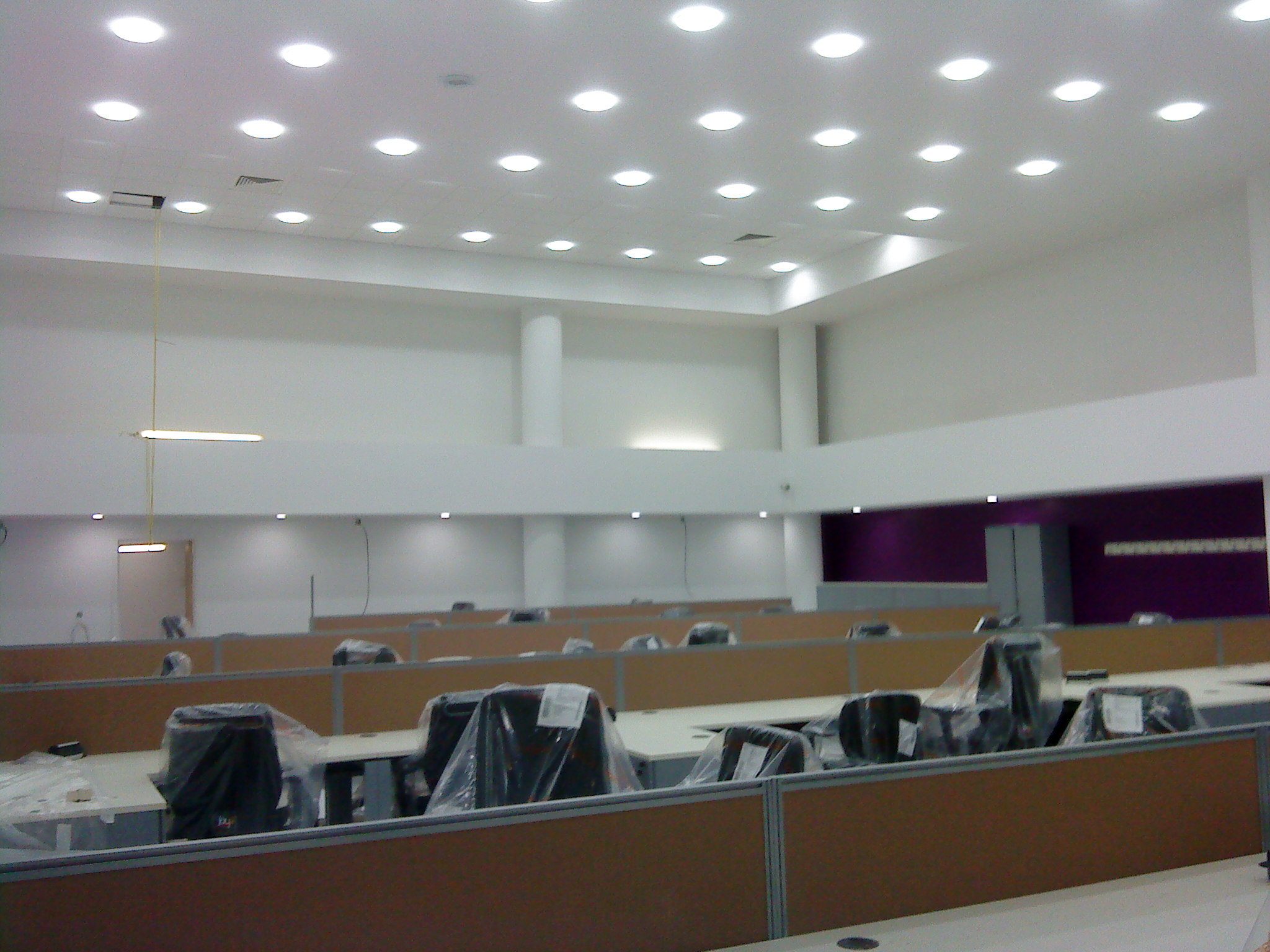 Nowe biuro typu open space z rzędami stanowisk pracy oddzielonych przegrodami, krzesła jeszcze w folii ochronnej, sufit z równomiernie rozmieszczonymi reflektorami.