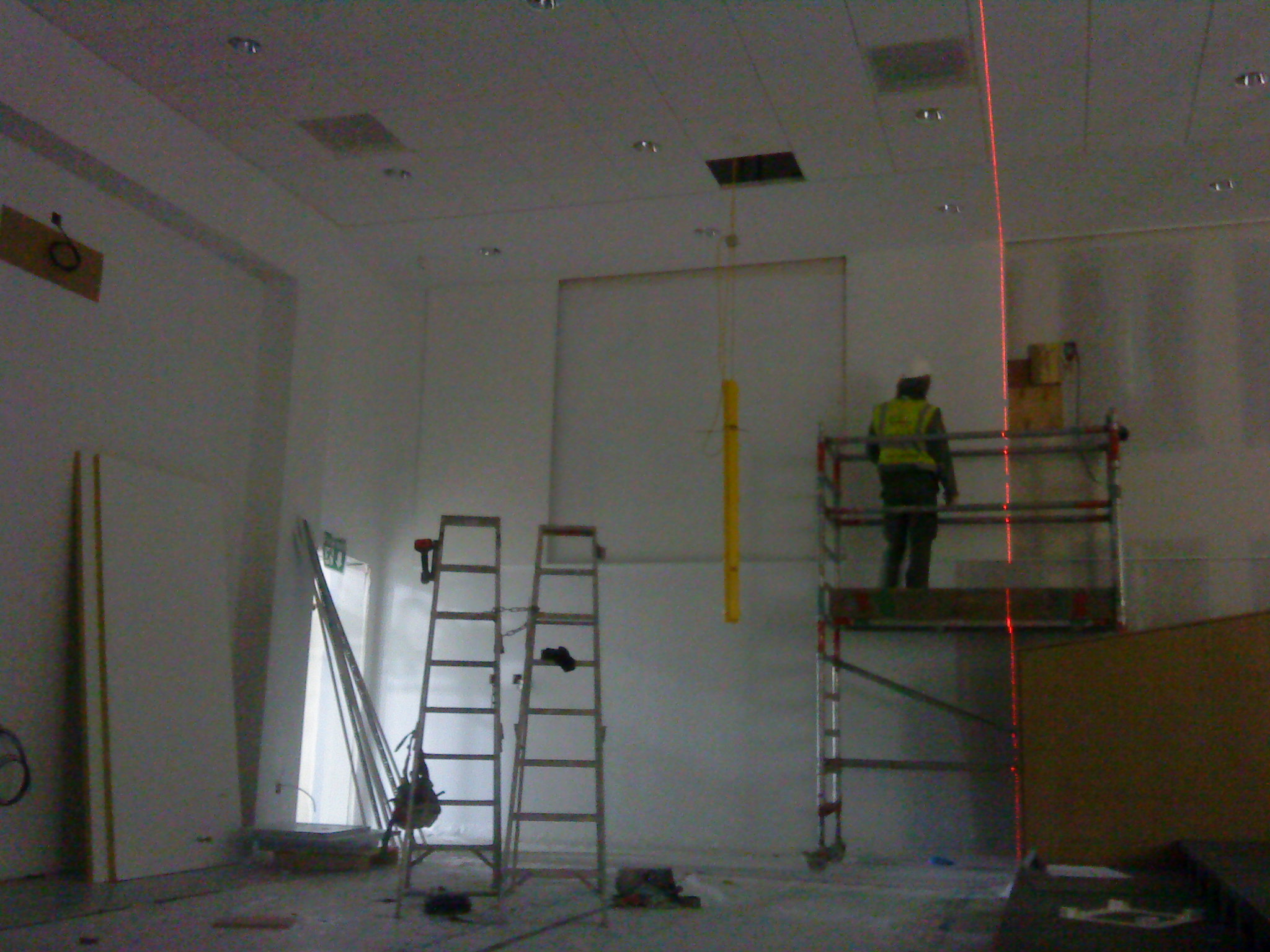 Wnętrze w trakcie wykończenia: pracownik w kamizelce na rusztowaniu, poziomica laserowa, drabiny, płyty gipsowe oparte o ścianę, surowy stan wykończenia.