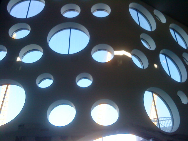 Ściana z licznymi okrągłymi otworami, przez które widać niebieskie niebo i fragment konstrukcji budynku, z widocznym żurawiem budowlanym w tle.