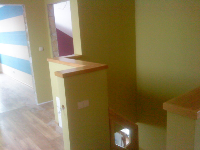 Wnętrze domu w trakcie wykończenia: pomalowane na zielono ściany, drewniane wykończenie schodów i podłoga, widoczne niedokończone prace przy ścianie w paski.