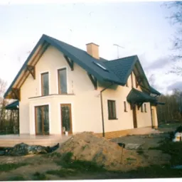 Jasny, dwukondygnacyjny dom jednorodzinny z ciemnym dachem i jasną elewacją, widoczny z perspektywy ogrodu z nasypem ziemi na pierwszym planie.