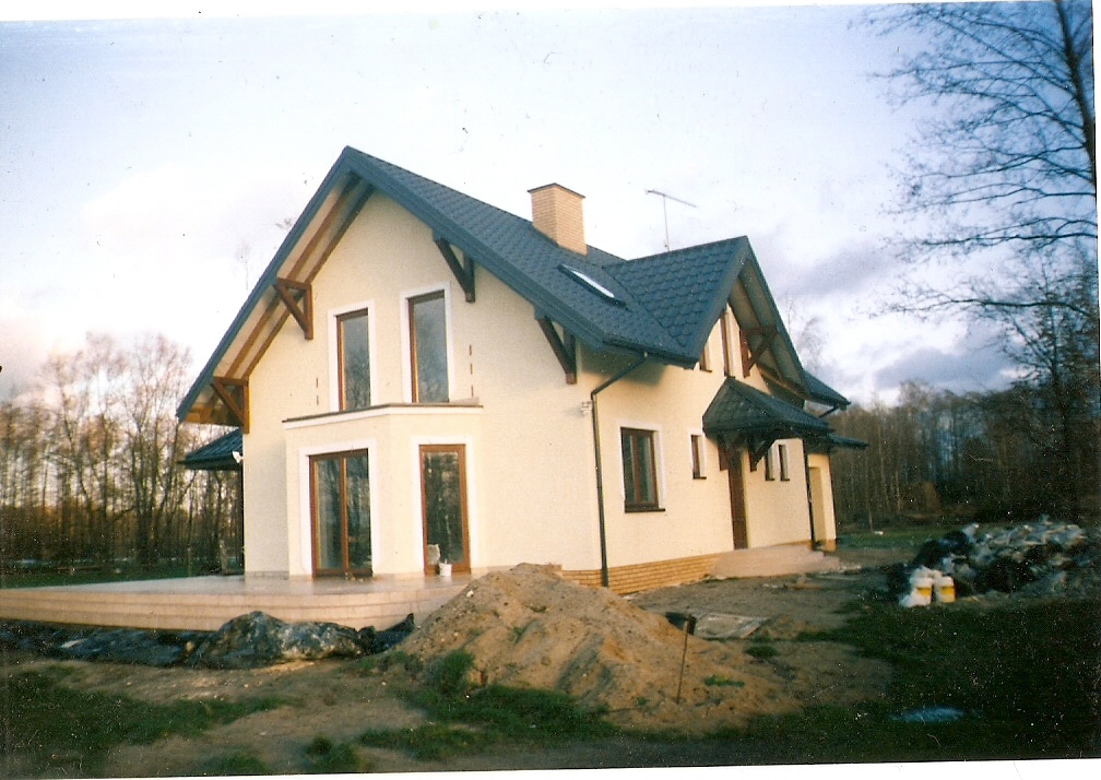 Jasny, dwukondygnacyjny dom jednorodzinny z ciemnym dachem i jasną elewacją, widoczny z perspektywy ogrodu z nasypem ziemi na pierwszym planie.