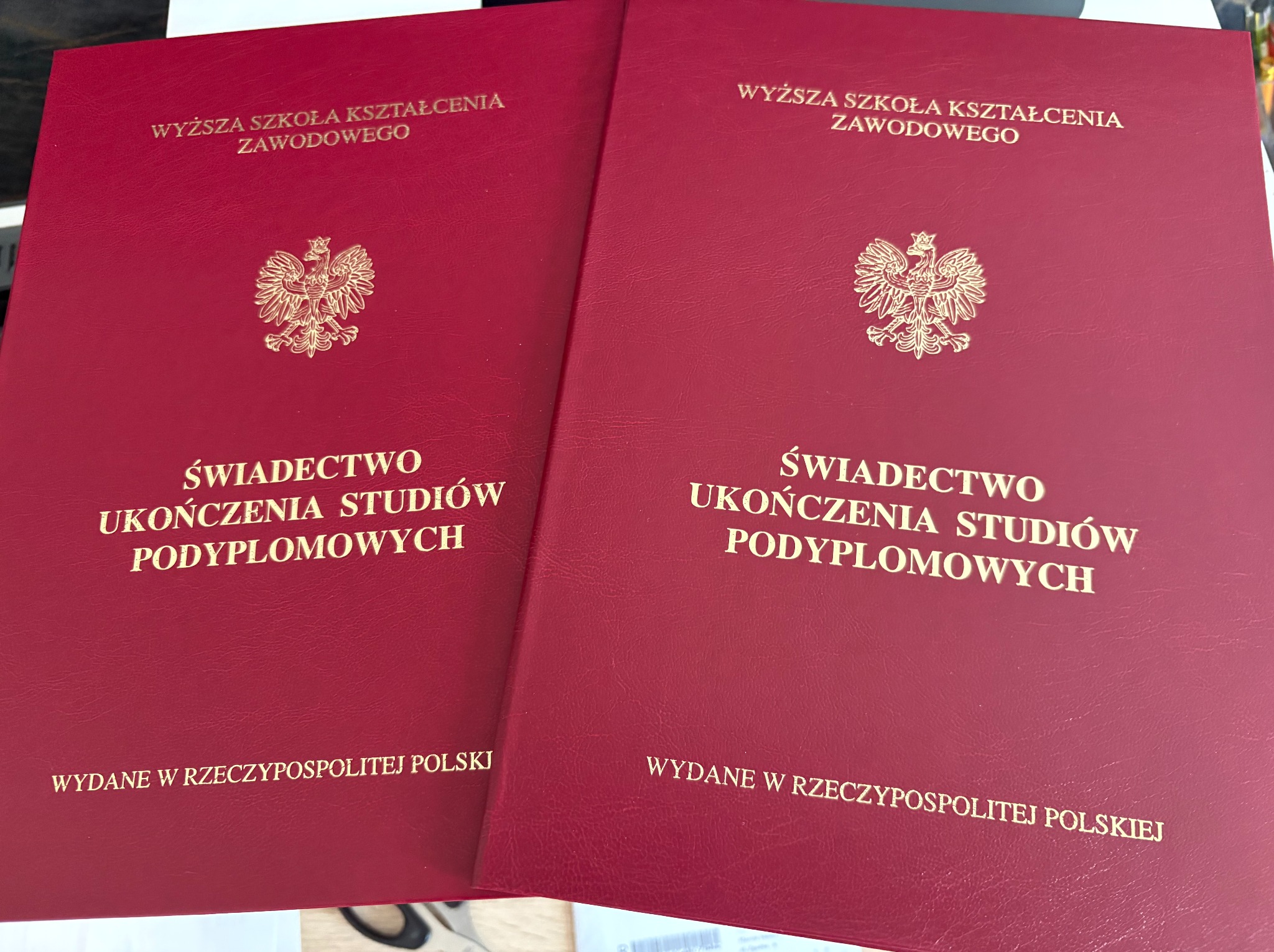 Dwa czerwone dyplomy ukończenia studiów podyplomowych Wyższej Szkoły Kształcenia Zawodowego z godłem Polski, wydane w Rzeczypospolitej Polskiej, leżą obok siebie.