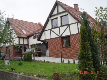 Dwukondygnacyjny dom w stylu szachulcowym z czerwonej cegły i białych belek, z garażem w bryle budynku, widok z zadbanego trawnika z młodymi tujami.