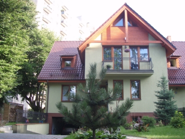 Dwukondygnacyjny dom jednorodzinny z poddaszem użytkowym, pokryty czerwoną dachówką, z jasną elewacją i drewnianymi elementami wykończeniowymi, otoczony zielenią, widoczny z poziomu podjazdu.