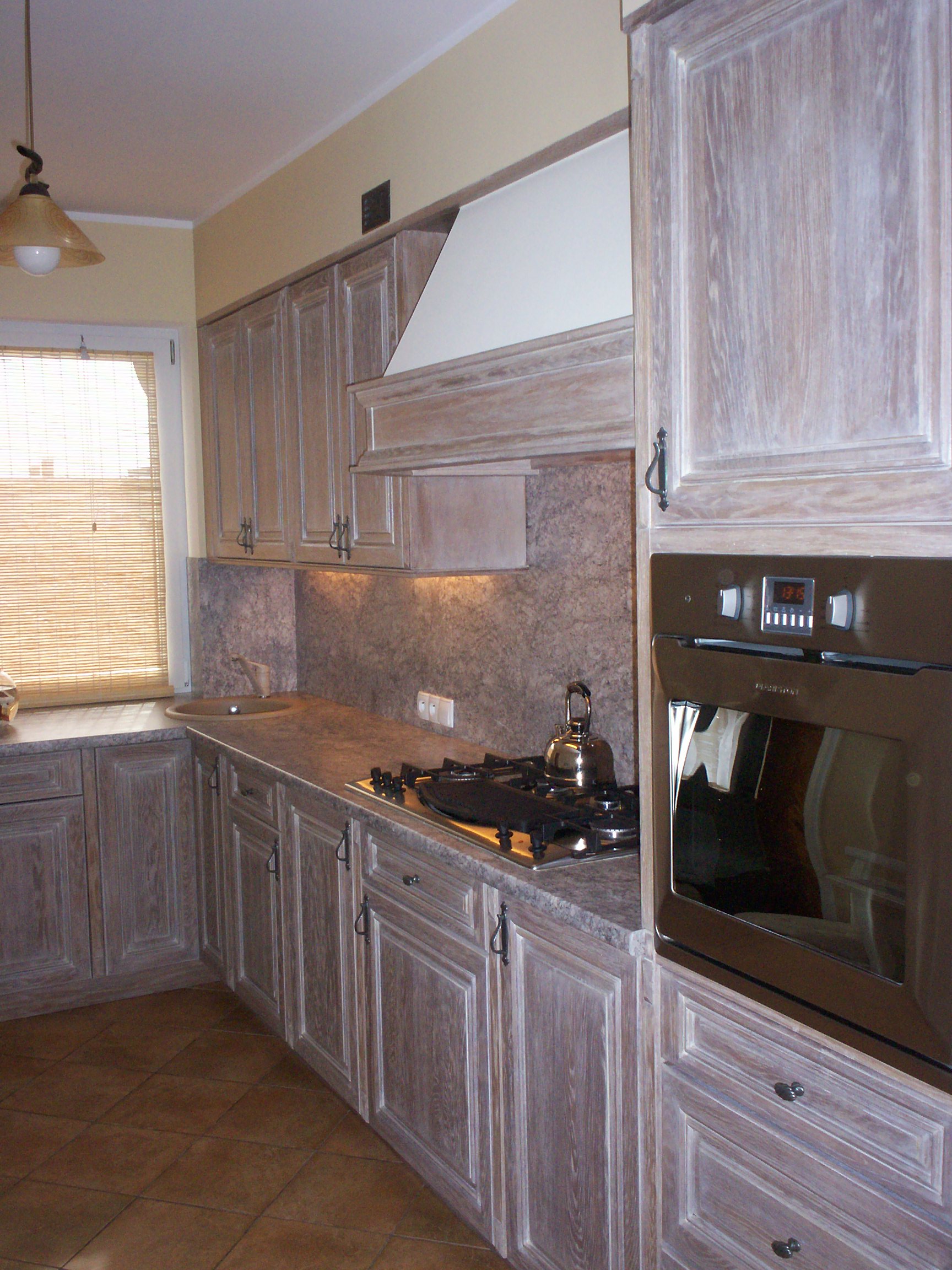 Jasna kuchnia z drewnianymi frontami, widoczny fragment blatu, płyty gazowej i piekarnika oraz brązowe płytki na podłodze.