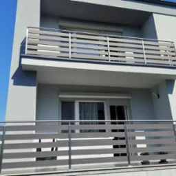 Nowoczesny balkon z poziomymi panelami w odcieniach szarości i srebrnymi słupkami, widok z dołu na tle błękitnego nieba.