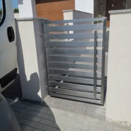 Nowoczesna, pozioma brama ze stali nierdzewnej, zamontowana pomiędzy dwoma słupkami z jasnego tynku, z widocznym fragmentem domu w tle.