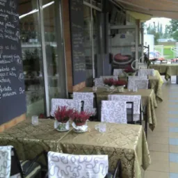 Ustawione stoły na zewnątrz restauracji z dekoracjami z wrzosów w doniczkach, nakryte złotymi obrusami i krzesłami z białymi pokrowcami z wzorem, menu wypisane kredą na tablicy w tle.