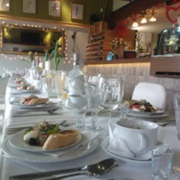 Uroczysty stół z białym obrusem, zastawiony talerzami z przekąskami, pieczywem, szklankami i kieliszkami, w tle dekoracje i butelki z napojami, aranżacja na przyjęcie rodzinne.