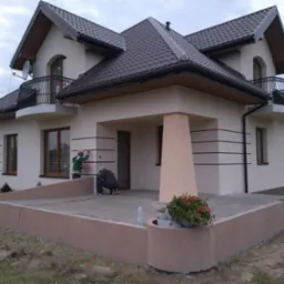 Elewacja domu jednorodzinnego w trakcie wykańczania, z widocznymi pracownikami przy montażu detali i wykończeniu tarasu. Kolorystyka: biel, brąz, beż.