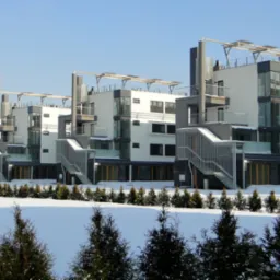 Szereg nowoczesnych, ocieplonych budynków mieszkalnych z płaskimi dachami i zewnętrznymi schodami w zimowym krajobrazie, widoczne ośnieżone trawniki i iglaki.