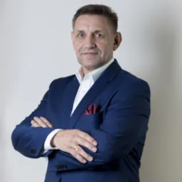 Mężczyzna w niebieskim garniturze i białej koszuli, z bordową poszetką w kieszeni, pozuje z założonymi rękami na neutralnym tle.