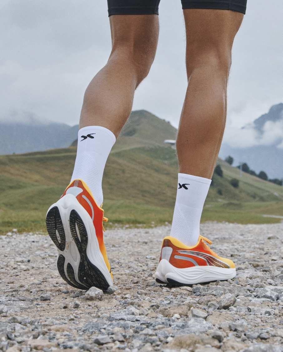 Biegacz w pomarańczowo-żółtych butach sportowych z białymi skarpetami z logo na kamienistej ścieżce górskiej