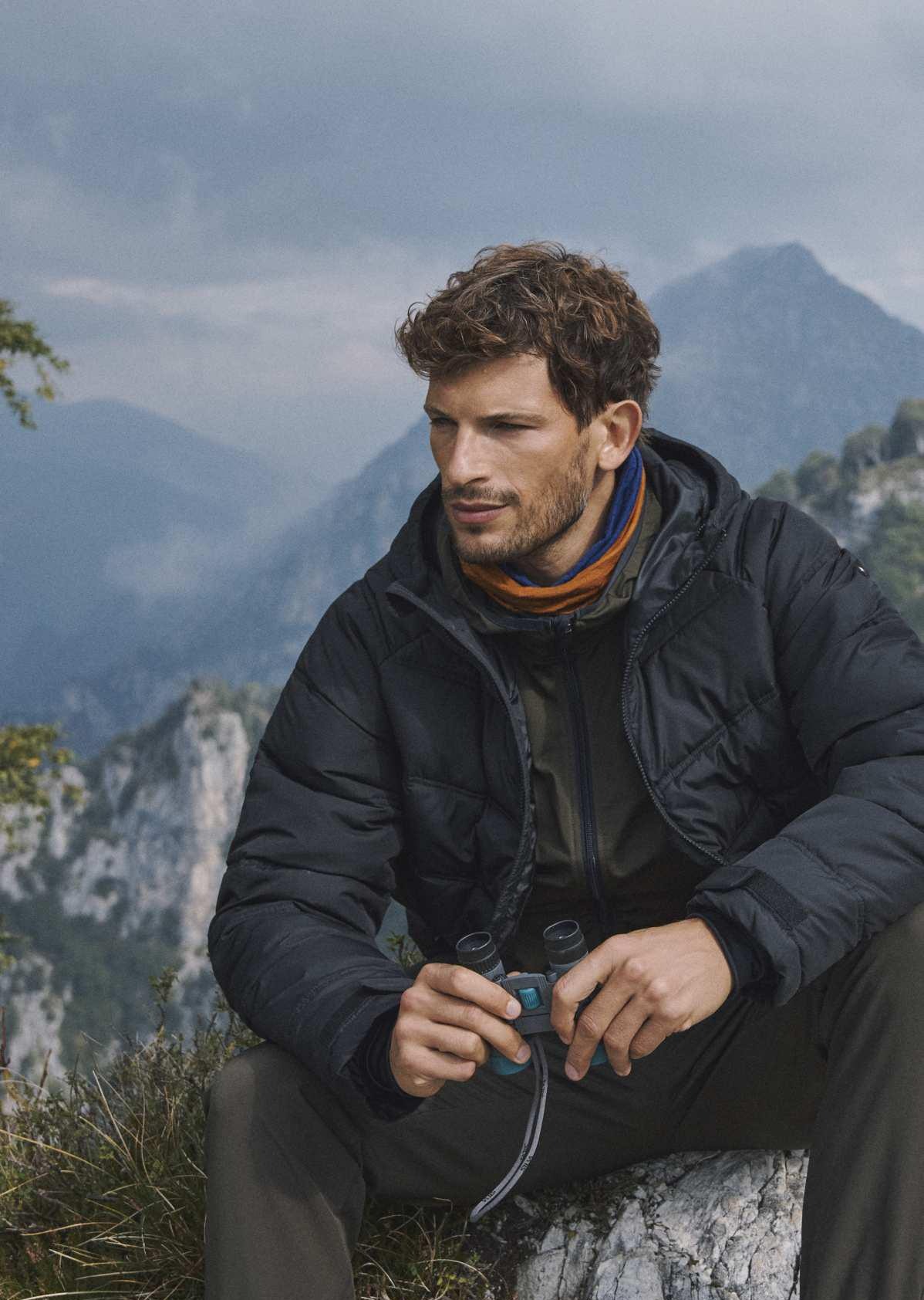 Mężczyzna z kręconymi włosami w czarnej kurtce puchowej, z lornetką w rękach, siedzi na tle górskiego krajobrazu.