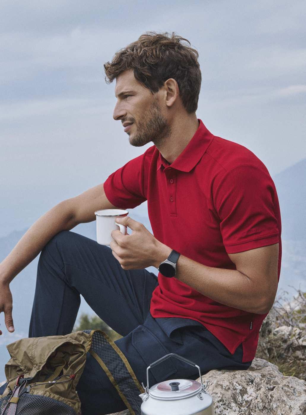 Mężczyzna w czerwonej koszulce polo siedzi na skale z kubkiem, w tle góry, obok plecak i czajnik.