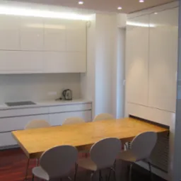 Nowoczesna, jasna kuchnia z białymi, błyszczącymi szafkami i drewnianym stołem z czterema krzesłami. Stół wysuwany spod zabudowy szafy.