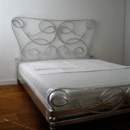 Metalowe łóżko z dekoracyjnym zagłówkiem w kształcie wijących się linii, ustawione na drewnianej podłodze przy białej ścianie.