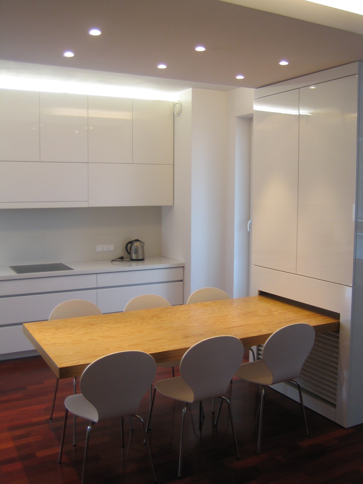 Nowoczesna, jasna kuchnia z białymi, błyszczącymi szafkami i drewnianym stołem z czterema krzesłami. Stół wysuwany spod zabudowy szafy.