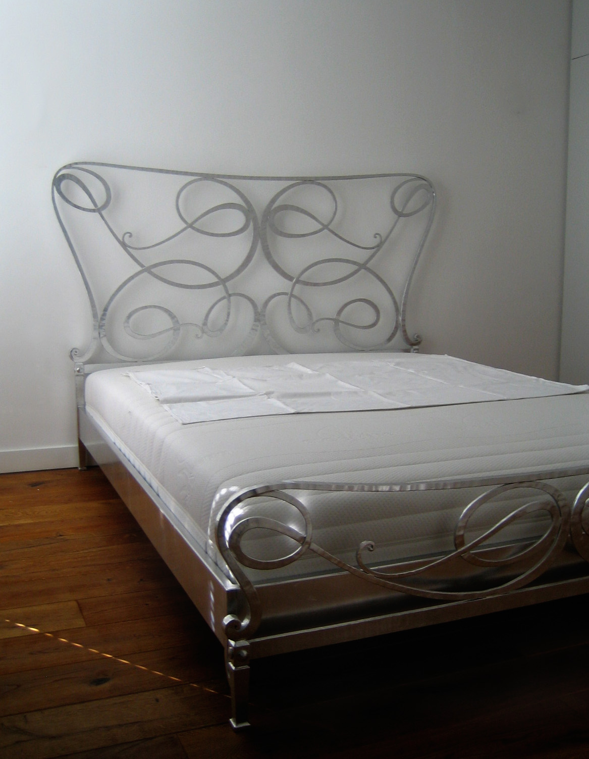 Metalowe łóżko z dekoracyjnym zagłówkiem w kształcie wijących się linii, ustawione na drewnianej podłodze przy białej ścianie.
