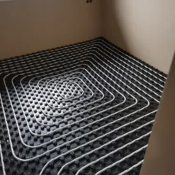 Białe rurki ogrzewania podłogowego ułożone spiralnie na czarnej macie izolacyjnej w jasnym pomieszczeniu z oknem.