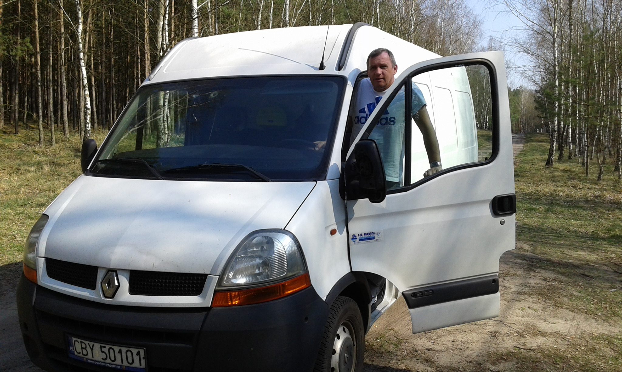Kierowca w białej koszulce, stojący w otwartych drzwiach białego busa Renault Master na tle lasu brzozowego, widoczna tablica rejestracyjna z Bydgoszczy.