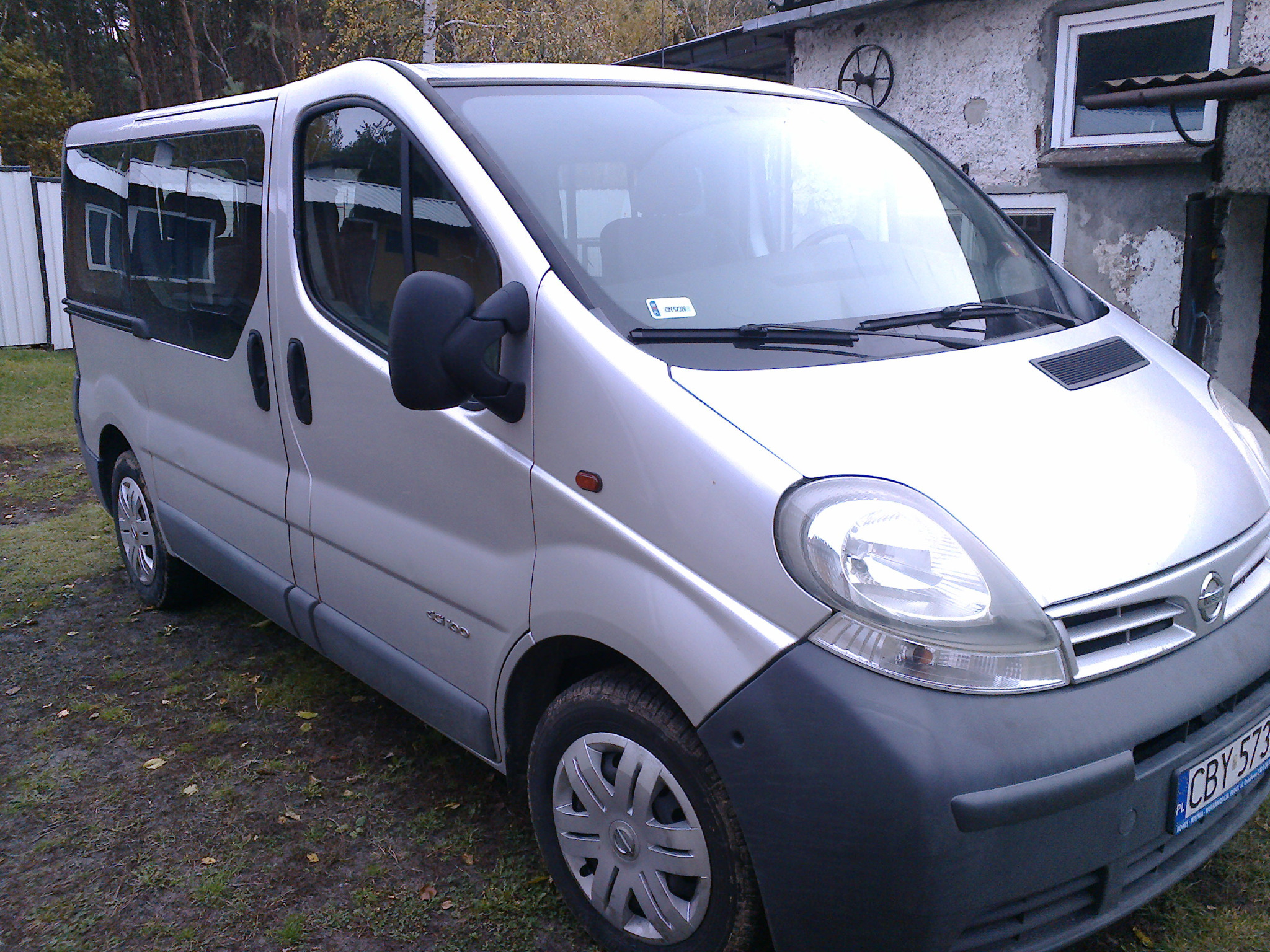 Srebrny bus Nissan z polskimi tablicami rejestracyjnymi zaparkowany na trawie, przed budynkiem otynkowanym na szaro. Widoczne drzewa w tle.