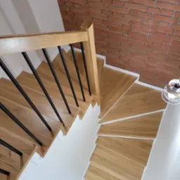 Kątowe ujęcie dębowych schodów z czarną balustradą, kontrastujące z ceglaną ścianą i białym wykończeniem, oświetlone wiszącą lampą.