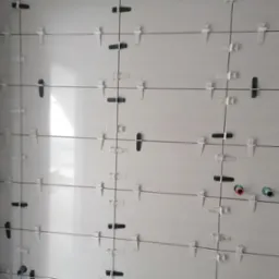 Świeżo ułożone białe płytki ceramiczne na ścianie, z widocznymi systemami poziomowania płytek w kolorze białym i czarnym oraz wystającymi rurami instalacyjnymi w kolorach czerwonym i niebieskim.