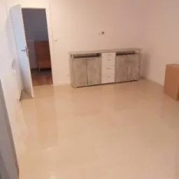 Wykończone wnętrze pokoju z jasną, błyszczącą podłogą z płytek, białe ściany i nowoczesna komoda z szarymi frontami, widok na otwarte drzwi do sąsiedniego pomieszczenia.