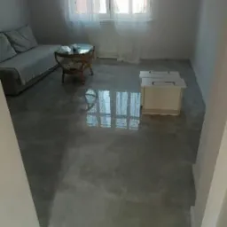 Wnętrze pokoju po wylaniu samopoziomującej wylewki, widoczny odblask okna w mokrej powierzchni, w tle sofa, stolik kawowy i grzejnik pod oknem.