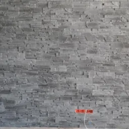 Ściana wykończona szarą okładziną kamienną, z widocznym gniazdkiem elektrycznym bez osprzętu i wystającymi przewodami.