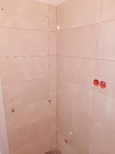 Ściana łazienki w trakcie wykańczania, widoczne jasne płytki ceramiczne imitujące marmur, z zaznaczonymi liniami wiercenia i otworami na instalacje, w tle ślady zaprawy.