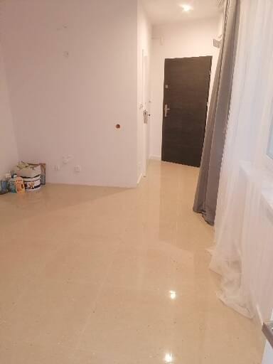 Puste pomieszczenie po remoncie z beżową podłogą, białe ściany z widocznymi śladami po gniazdkach i puszkach, ciemne drzwi wejściowe i materiały budowlane w rogu.