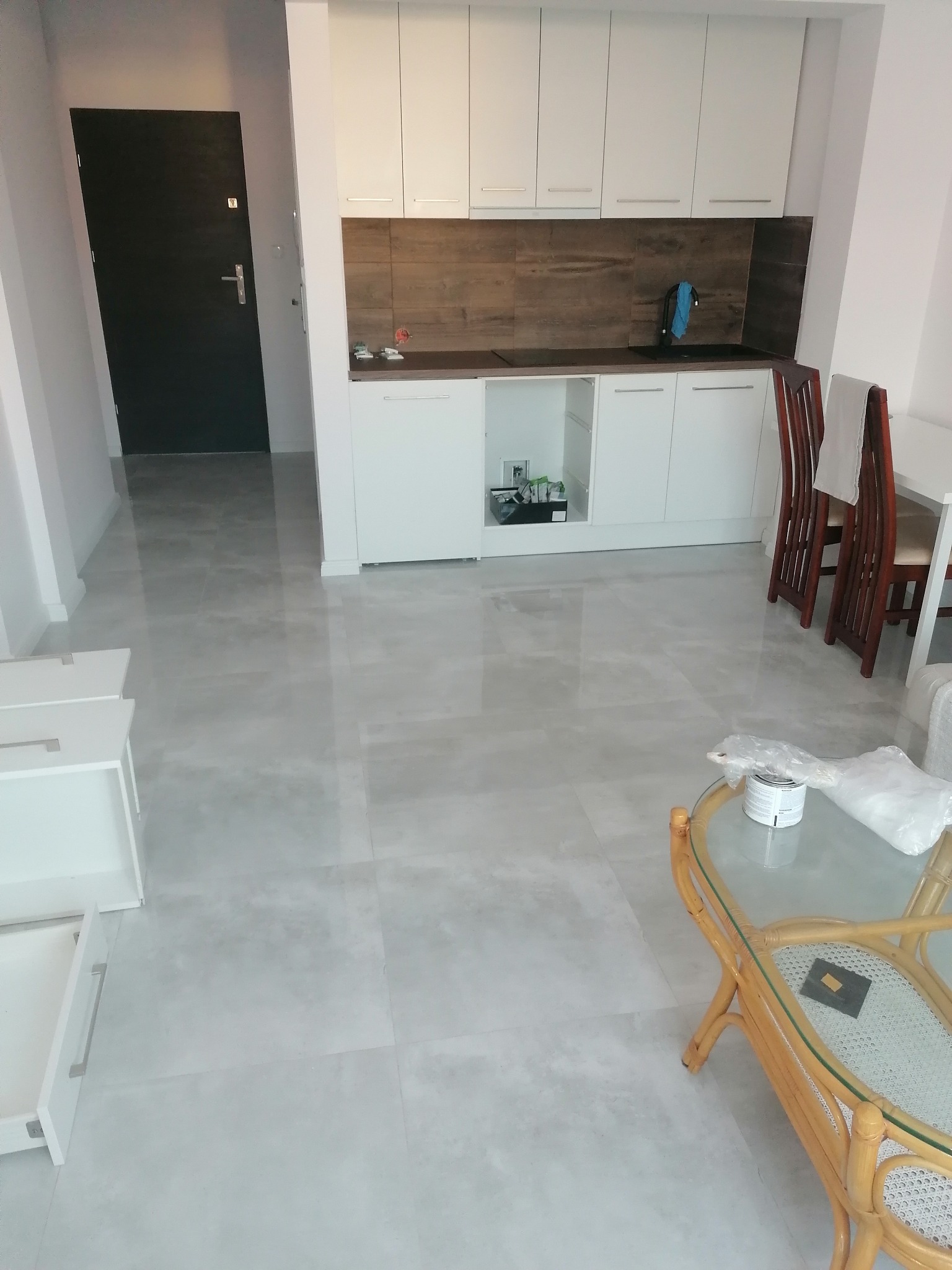 Jasne wnętrze mieszkania po remoncie: białe szafki kuchenne z drewnianym panelem nad blatem, szare płytki podłogowe, częściowo widoczne meble w stylu rattanowym i stół z krzesłami. Puszka z farbą...