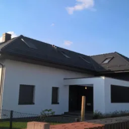 Nowoczesny dom jednorodzinny z ciemną dachówką i jasną elewacją, widoczne okna dachowe, fragment ceglanego ogrodzenia i błękitne niebo.