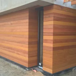 Fragment elewacji budynku wykończony poziomymi panelami z drewna egzotycznego, widoczny betonowy element konstrukcyjny i okno zabezpieczone folią malarską.