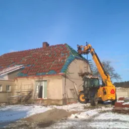 Dom z częściowo zdjętym, czerwonym dachem, robotnicy na podnośniku układają nowe dachówki, w tle błękitne niebo i śnieg na ziemi.