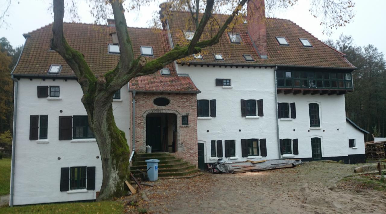 Rozległy, biały dom z ciemnymi okiennicami i czerwoną dachówką, widoczny z zewnątrz, z drzewem na pierwszym planie i materiałami budowlanymi na ziemi.