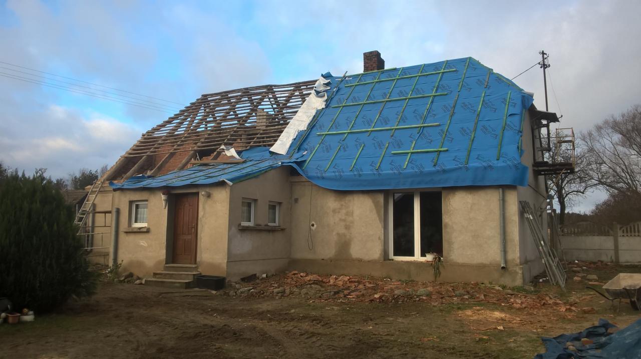 Dom w trakcie remontu dachu, połowa konstrukcji odsłonięta, druga połowa zabezpieczona niebieską plandeką, widoczne rusztowania i gruz.